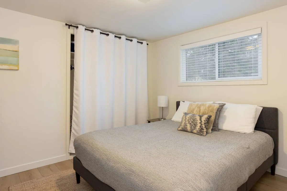 A serene bedroom featuring a queen-sized bed with gray bedding, decorative pillow, white curtains softly filtering natural light, and a bedside lamp. The room exudes calm and comfort, suggestive of restful nights and peaceful mornings.