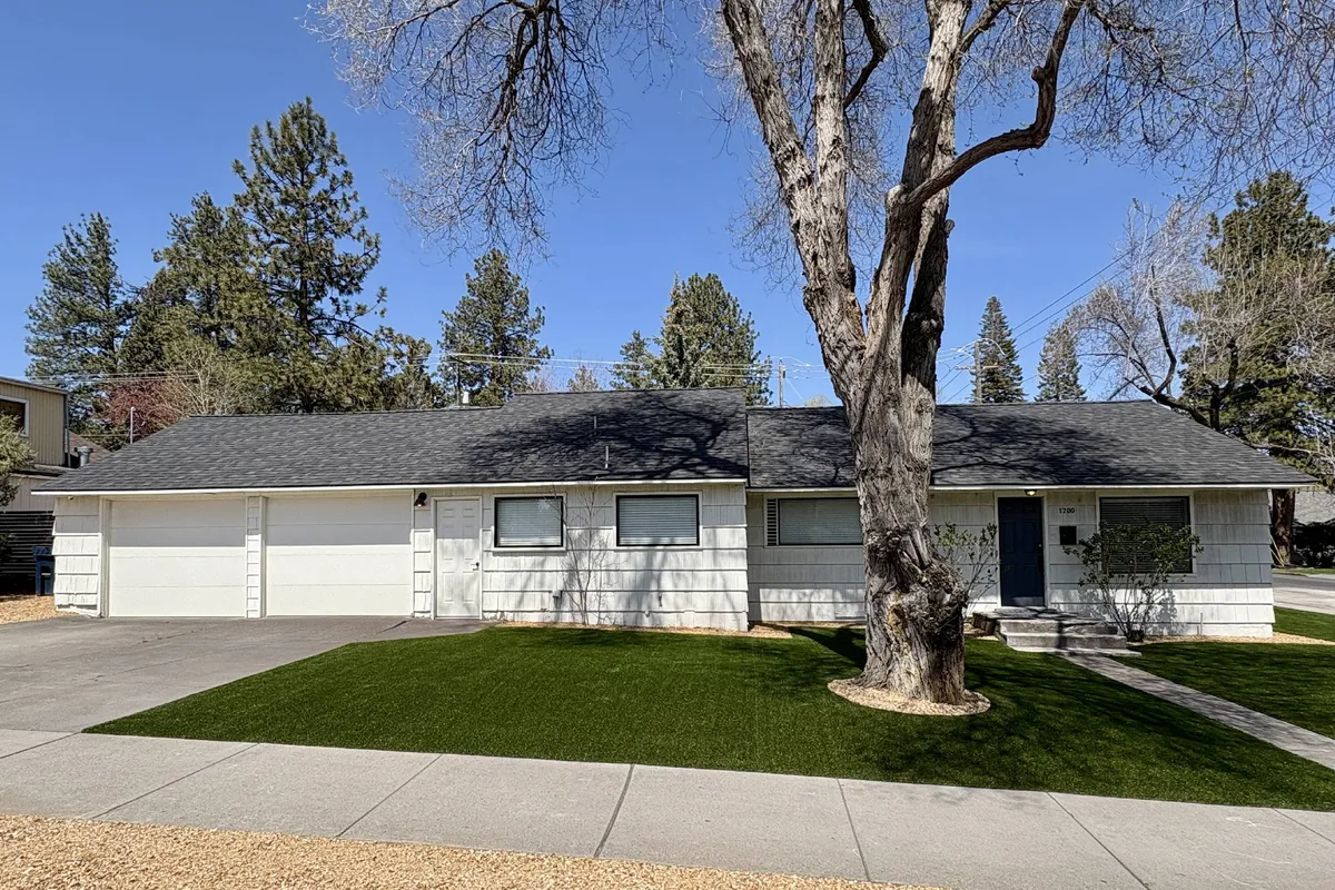 The image shows the welcoming exterior of a single-family home with a charming main entrance, a large mature tree providing shade, a lush, well-maintained lawn, and a spacious driveway. The scene suggests a perfect setting for outdoor gatherings, family barbecues, or relaxing summer evenings, all set in a vibrant Bend neighborhood.