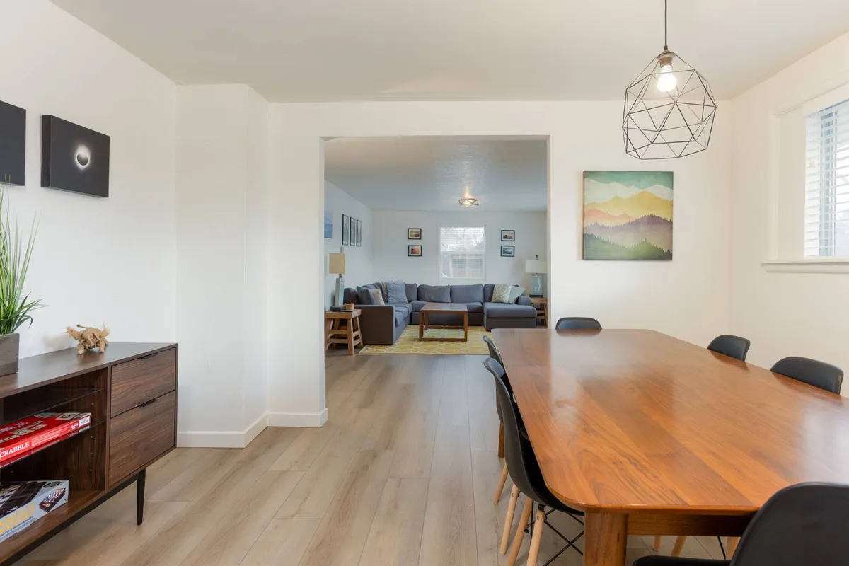 A bright view of the open-concept dining area with a table set for six, a striking pendant light above, and a glimpse into the adjoining living room. The space is filled with natural light, emphasizing a welcoming environment for family dinners and group gatherings.