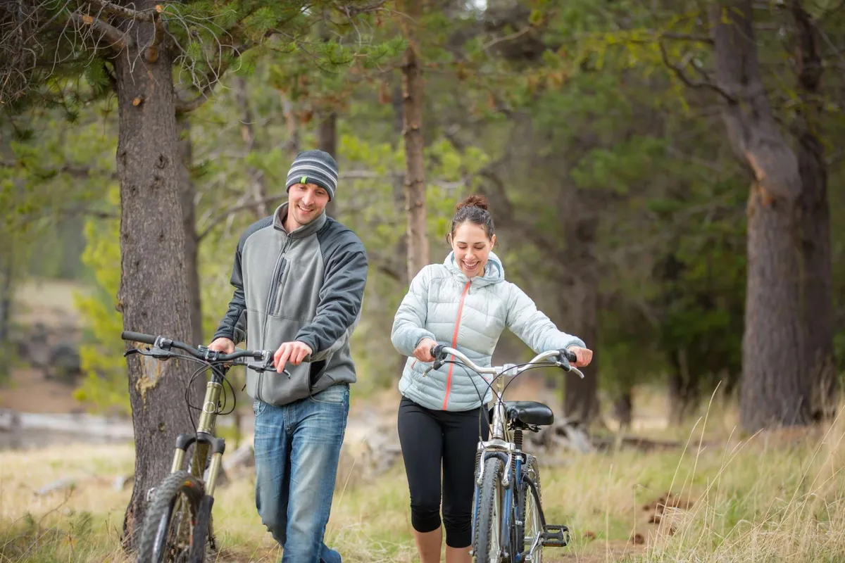 This image shows two people walking their bicycles along a grassy path framed by tall trees, evoking the experience of exploring nearby trails or biking through forested routes. The mood is joyful and adventurous.