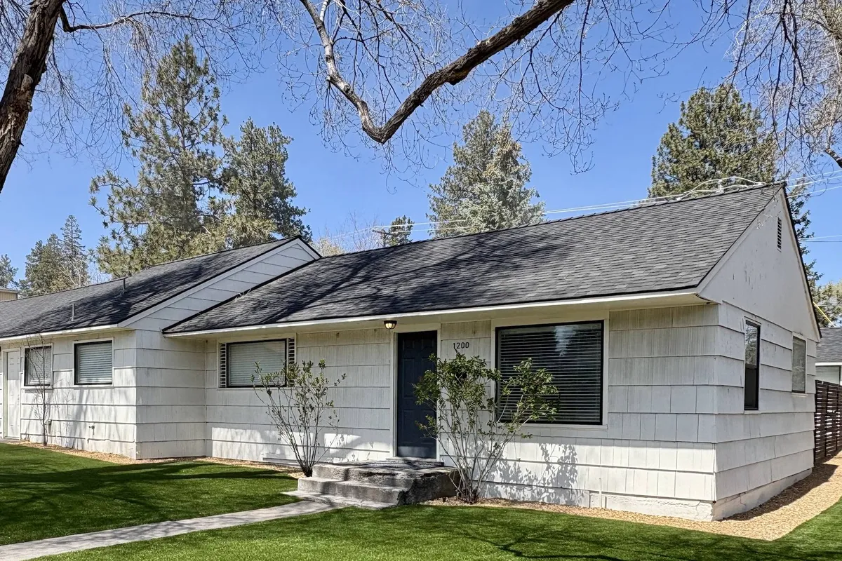 The photo shows a lush, landscaped lawn with the front door of the home visible, shaded by tree branches. People are relaxing in the yard, evoking a sense of relaxation and family-friendly gathering space.