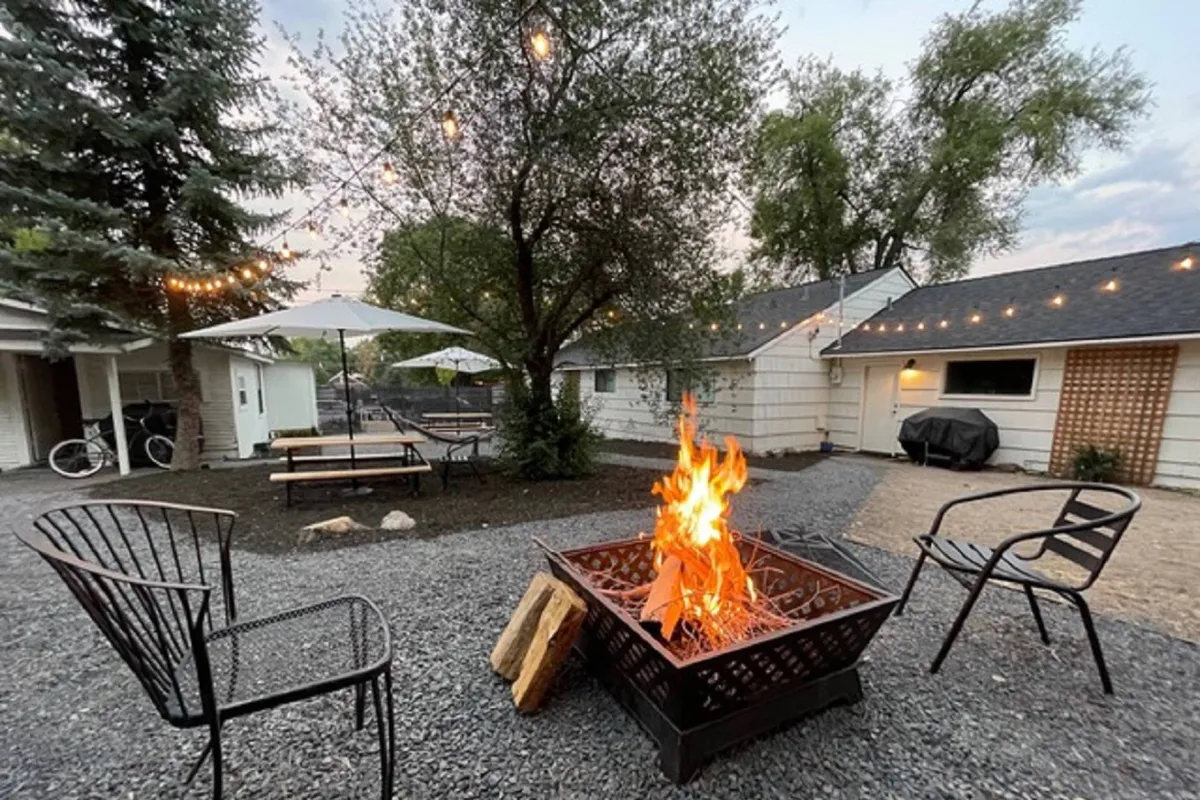 This image captures a glowing fire pit surrounded by outdoor seating under string lights, set amidst mature trees. A group gathers around, enjoying the inviting warmth, with a welcoming, communal atmosphere evident.