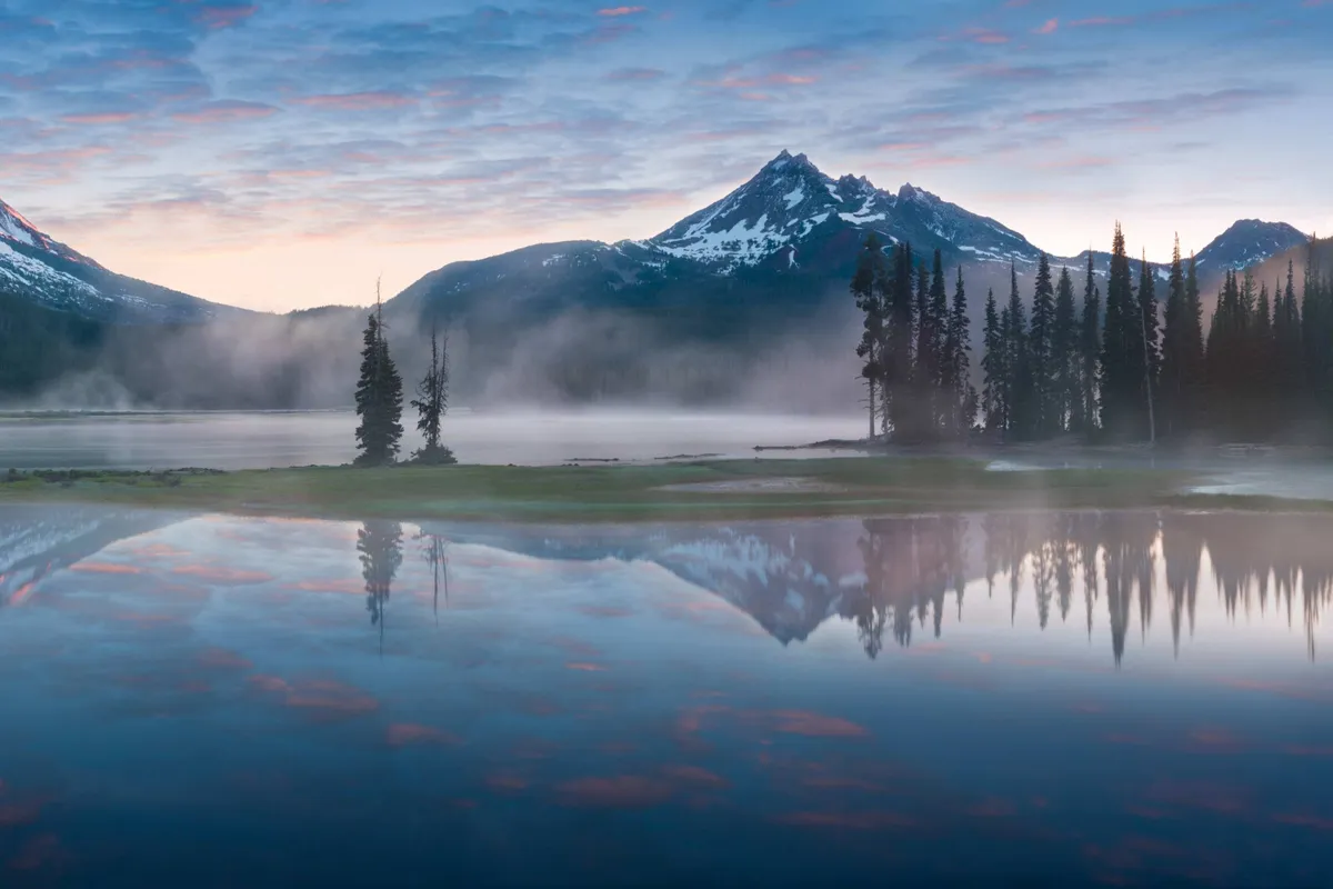 A tranquil lakeside scene with majestic mountains rising in the background, framed by tall trees and a wide sky. The open natural setting embodies the freedom and adventure of Bend, suggesting group outings and exploration beyond the home.