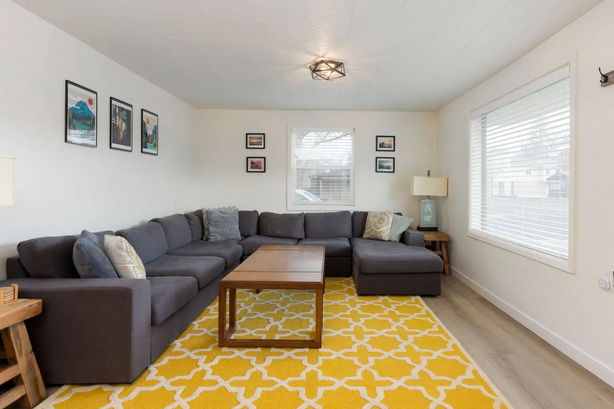 A welcoming living room with a large sectional sofa, bright yellow rug, wooden coffee table, and framed photos on the wall—offering space for group gatherings, casual conversations, and cozy evenings together.
