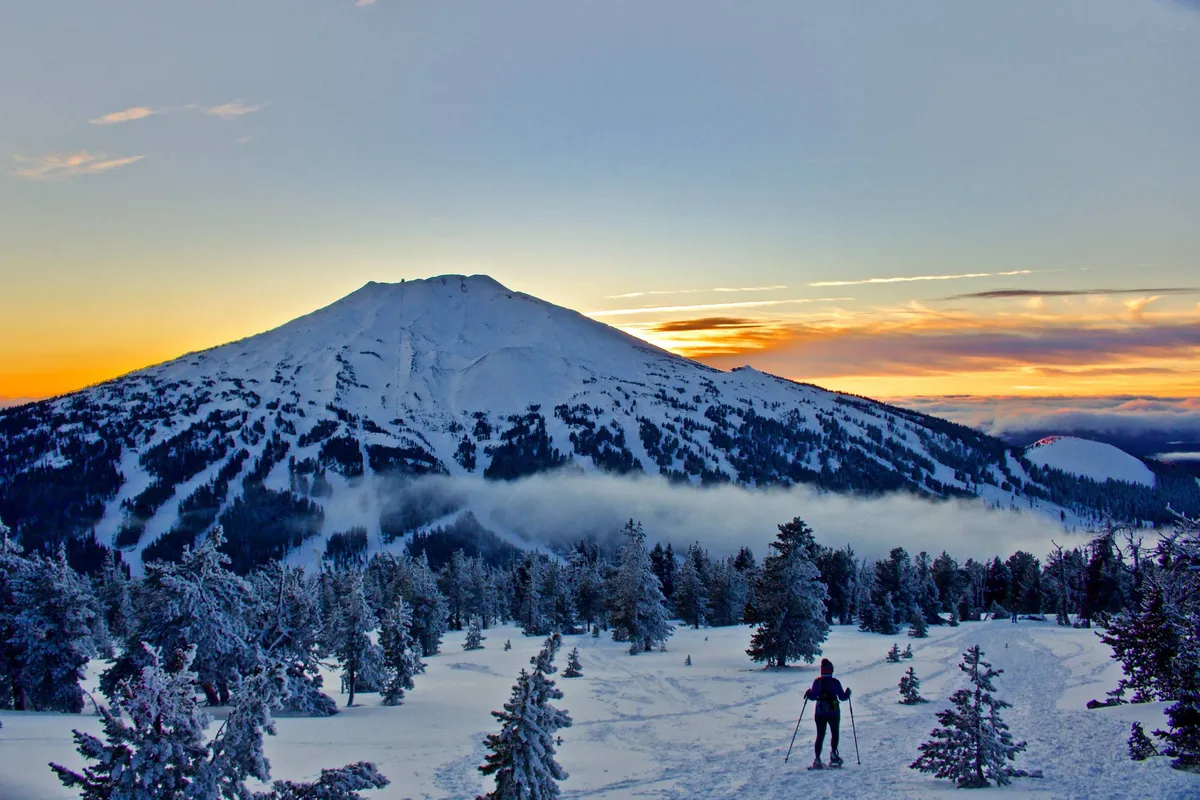 A striking scene of a snow-covered mountain at sunset, with a person skiing down the slope. The warm colors of the sky contrast with the cool tones of the snow, creating a dynamic and inviting atmosphere.